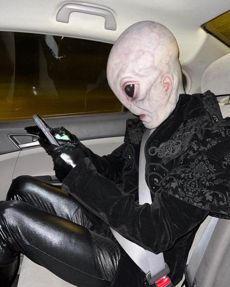 Wearing the Alien Area 51 mask and full outfit, checking a phone in the backseat of a car on the way to a Halloween party — The Terror Lab.