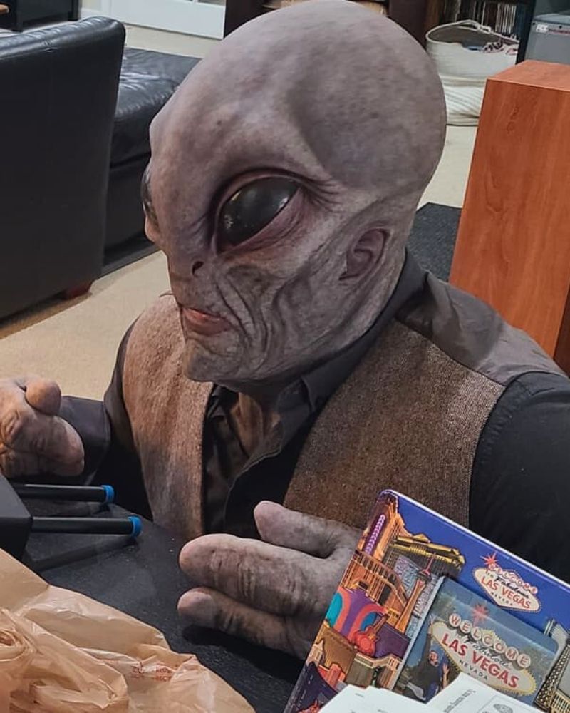 Wearing the Alien Area 51 realistic mask and gloves, sitting at a desk in a Las Vegas shop—looking like negotiations with humans have just begun — The Terror Lab.