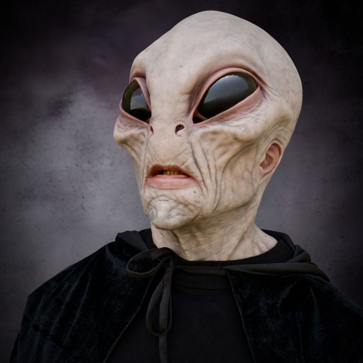 Alien creature mask with large eyes and a black cloak against a dark background