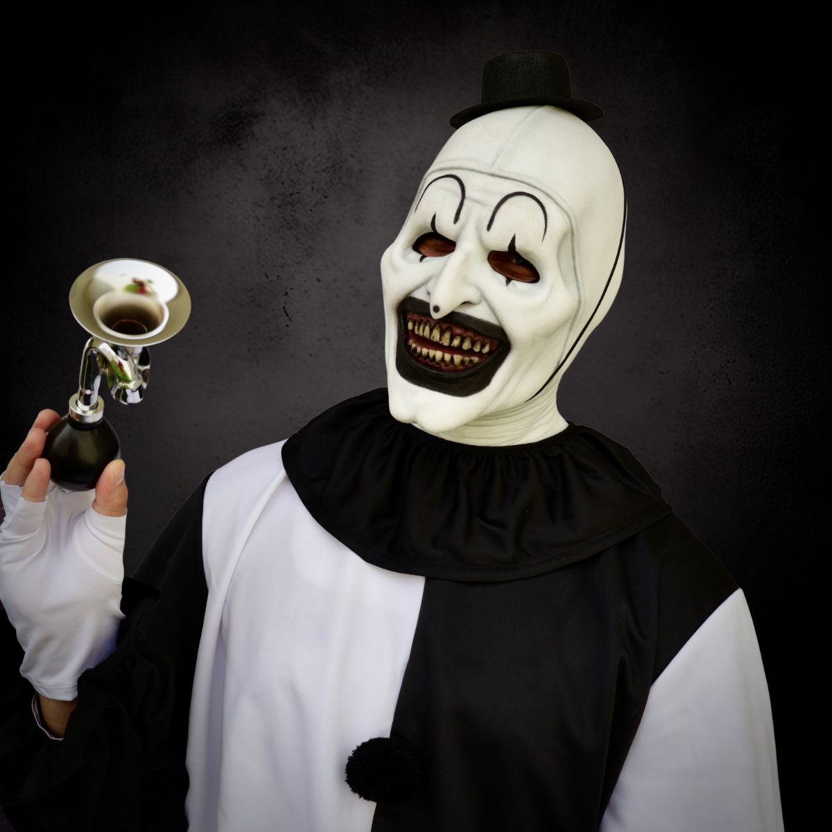 Person wearing Art the clown white mask with black accents, holding a horn with a white top against a dark background.