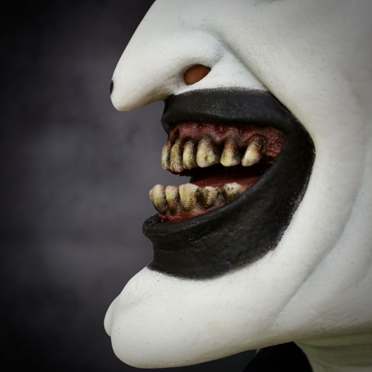 Close-up of art the clown halloween mask with a white face, black beard, and detailed teeth against a dark background.