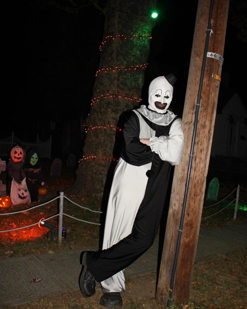 Wearing the Art the Clown mask and full costume, leaning casually against a wooden pole in front of a decorated Halloween yard — The Terror Lab.