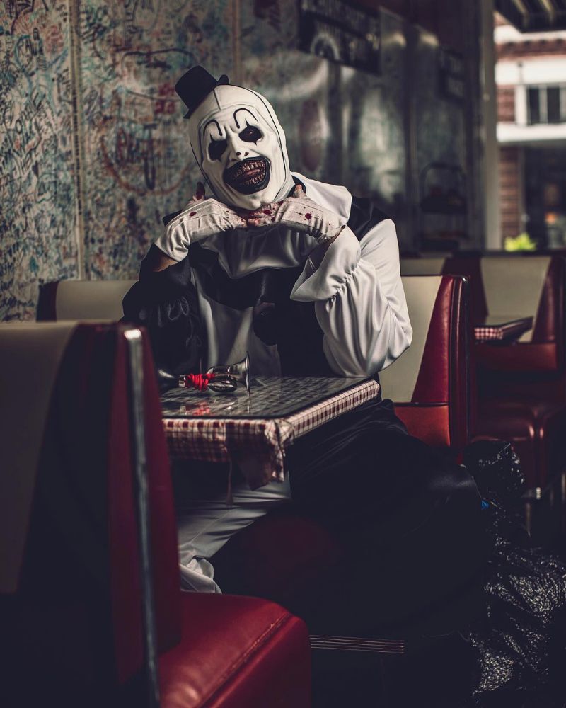 Art the Clown sitting in a retro diner, hands under chin and blood on gloves, during a Halloween night — The Terror Lab.