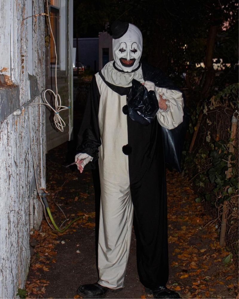 Wearing the Art the Clown mask and full costume, walking through a narrow backyard path with a trash bag during a Halloween night — The Terror Lab.