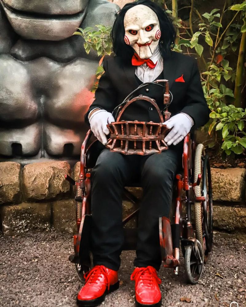 Customer wearing the Billy the Puppet moving jaw mask and full costume, seated in a distressed wheelchair holding a replica reverse bear trap — from The Terror Lab