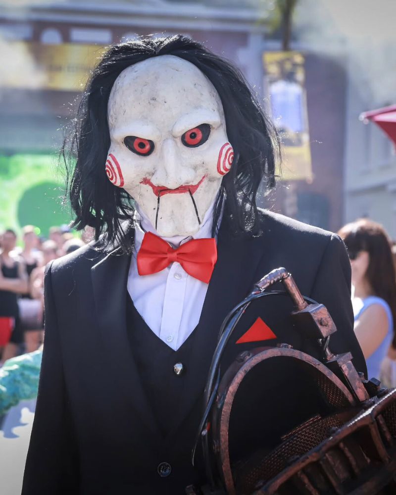Customer wearing the Billy the Puppet moving jaw mask with full tuxedo costume and red bow tie, holding the reverse bear trap prop during a crowded Halloween event — The Terror Lab.