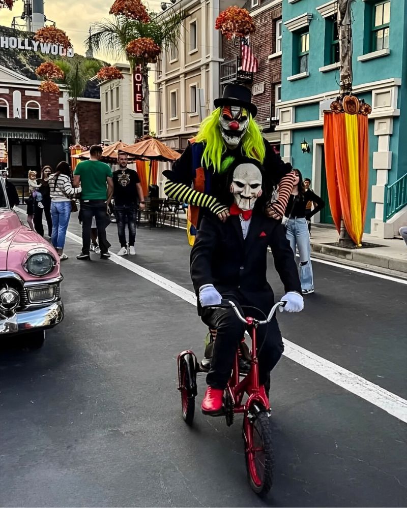 Customer dressed as Billy the Puppet with moving jaw mask, black suit and red shoes, riding a red tricycle at a Halloween street event, with a scary clown standing behind — from The Terror Lab.