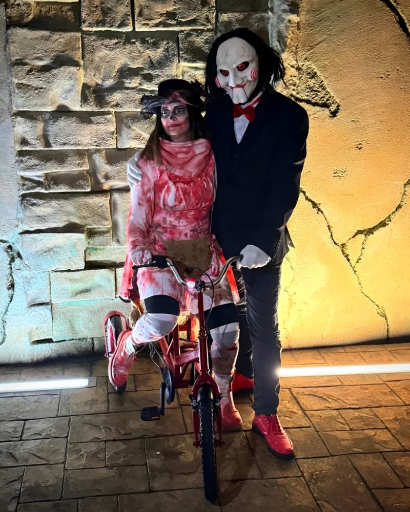 Customer in Billy the Puppet moving jaw mask posing with a woman in bloody bandage costume on a red tricycle during a Halloween night event — The Terror Lab.