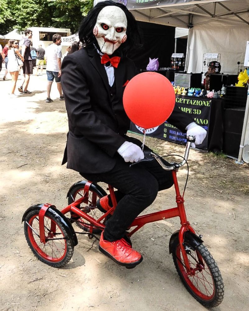 Customer riding a red tricycle while wearing the Billy the Puppet moving jaw mask and holding a red balloon at an outdoor Halloween event — from The Terror Lab