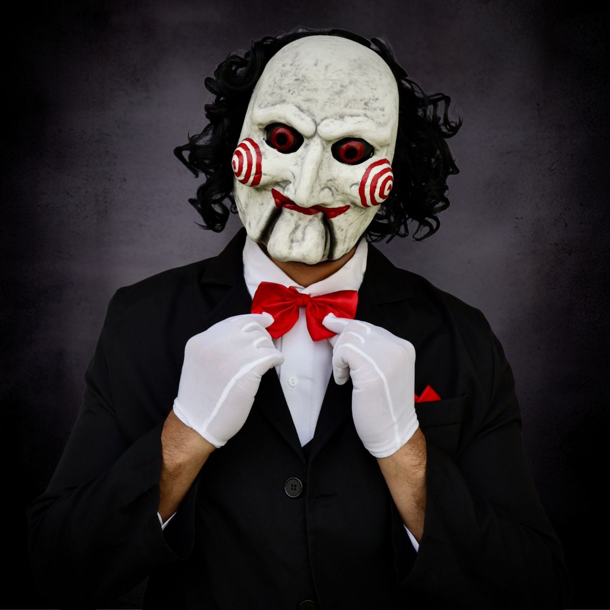 Person wearing horror white mask with red accents, black suit, and red bow tie against a dark background