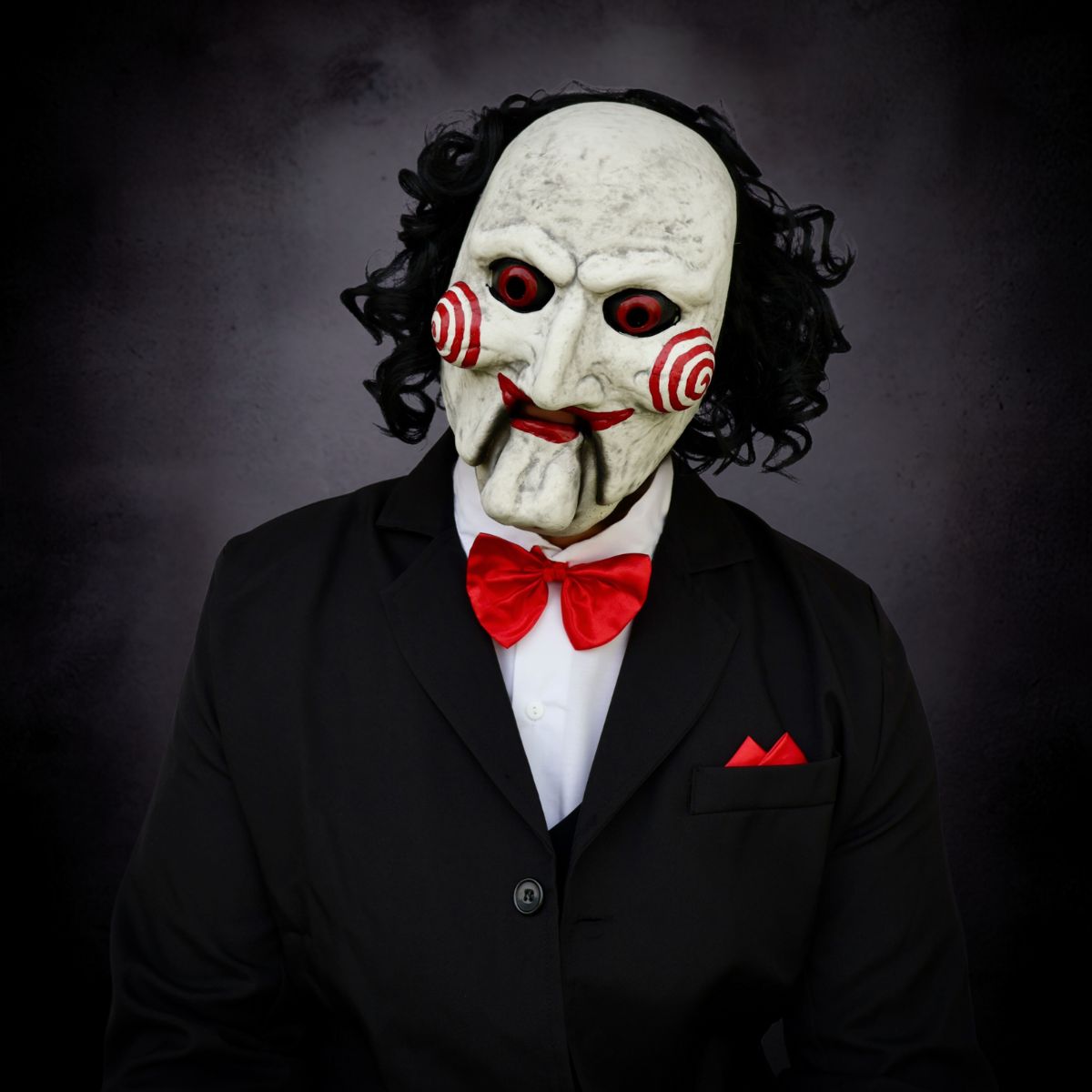 Person wearing a white mask with red eyes and a red bow tie against a dark background