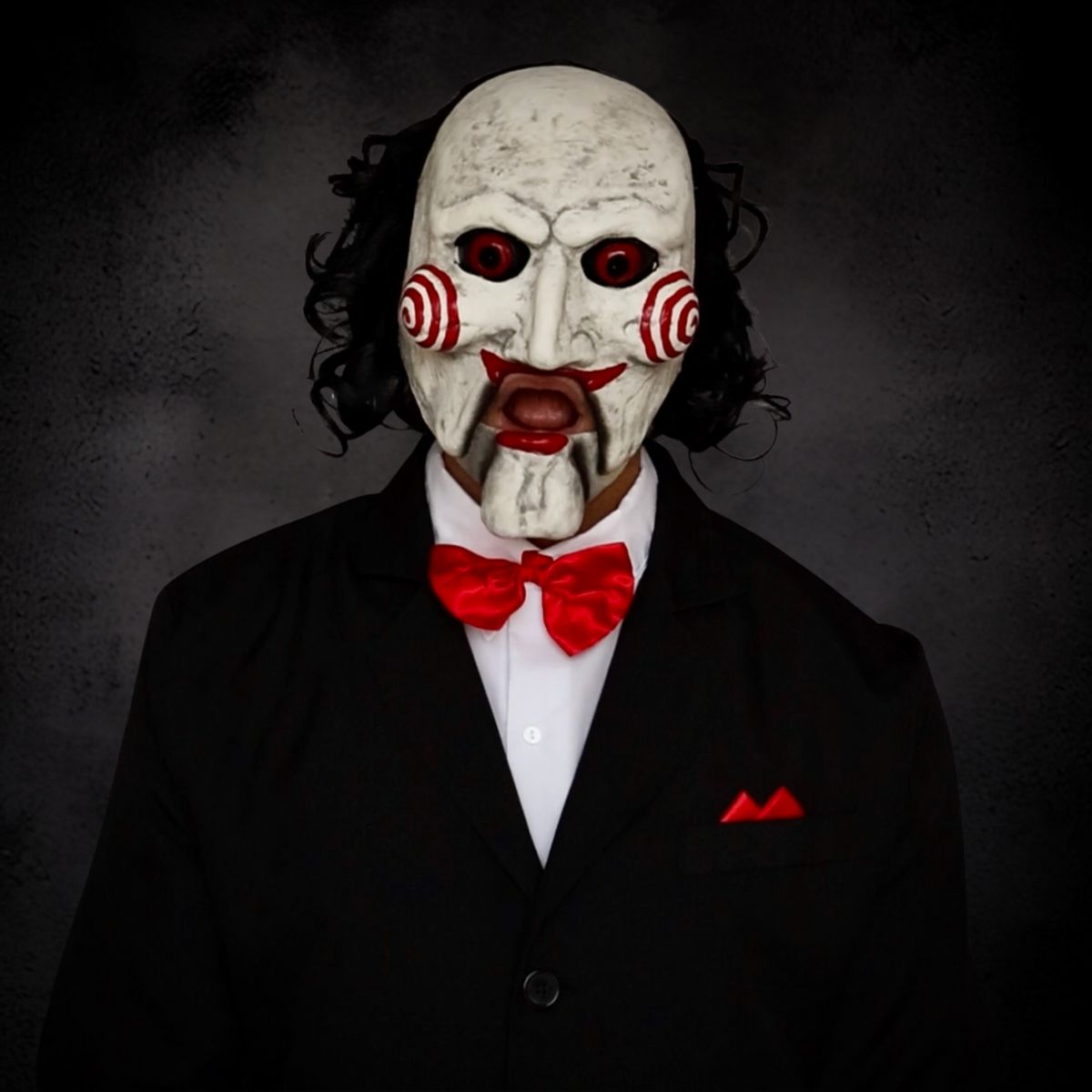Person wearing billy puppet white mask with red eyes, open mouth and a red bow tie, standing against a dark background.