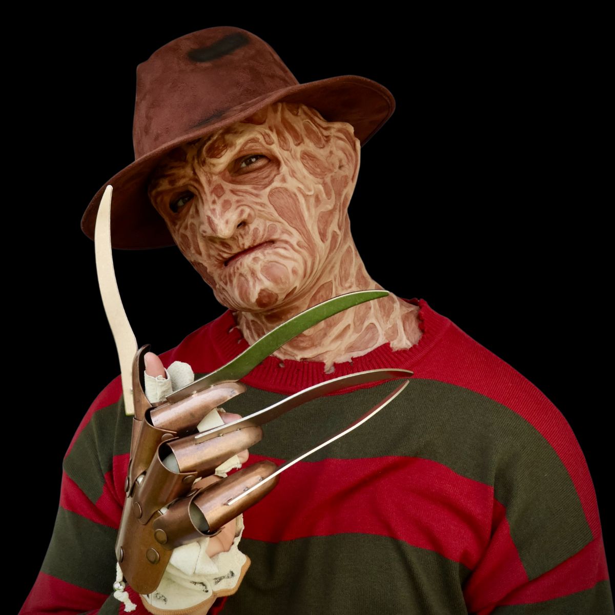 Freddy Character Halloween mask in a red and green striped shirt with a brown hat holding a claw glove against a black background