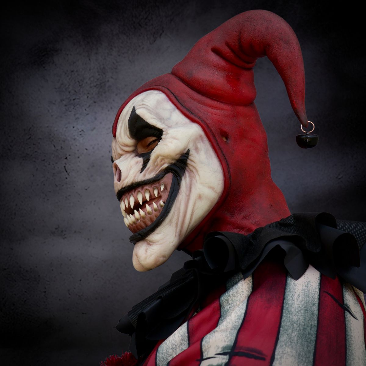 Horror-themed jester clown mask with red hood and white face on a dark background