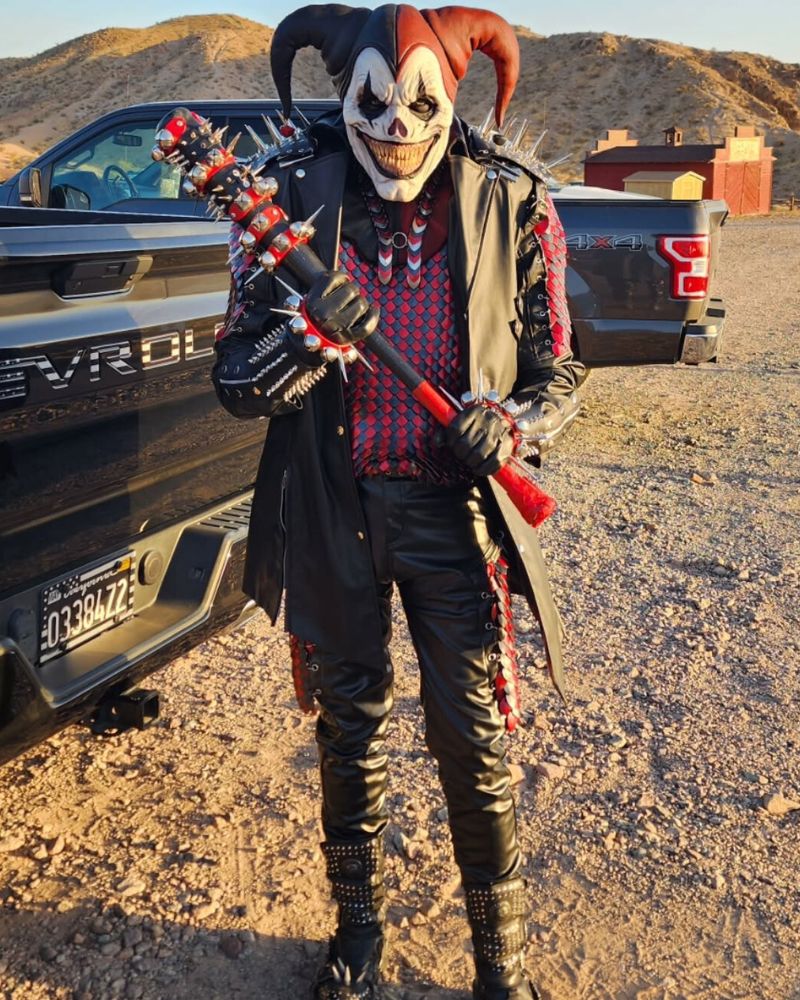 Jester clown mask paired with black and red leather outfit, holding a spiked bat in the desert at golden hour — The Terror Lab.