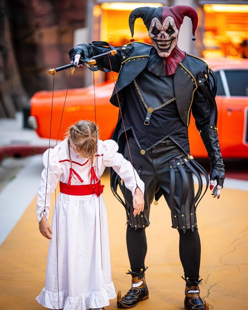Red and black jester realistic mask paired with a sinister marionette setup on Halloween night — striking street performance captured outside a haunted attraction. The Terror Lab.