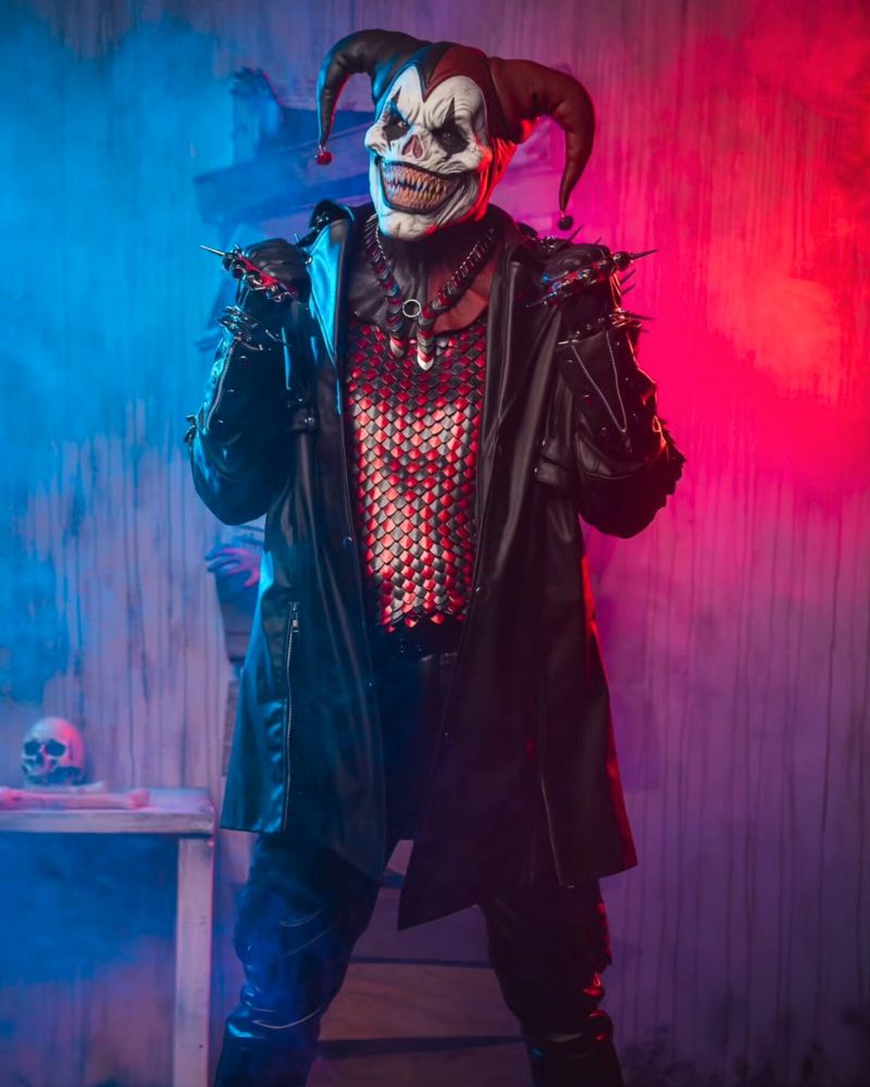 Sinister jester clown mask worn with full leather outfit and red mesh armor, captured in a smoke-filled haunted set with red and blue lighting — The Terror Lab.