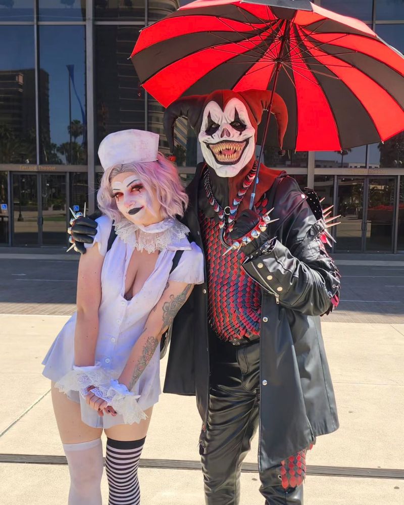 Jester clown mask paired with full leather and red mesh costume, posing outside a convention with a gothic nurse character under a red-and-black umbrella — The Terror Lab.