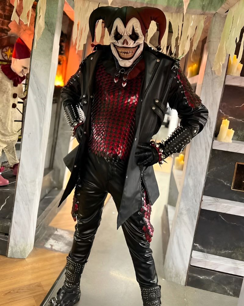 Evil jester mask worn with spiked leather outfit and red scale armor, posing confidently inside a haunted house set with candles and Halloween props — The Terror Lab.