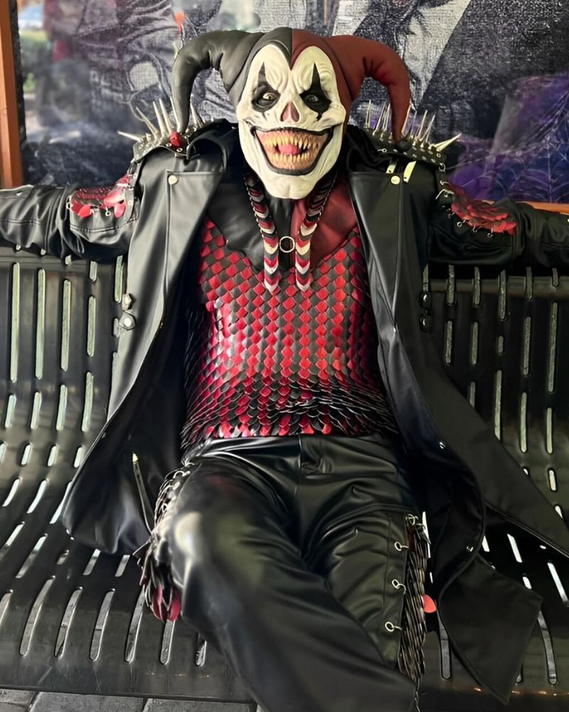 Jester mask with sinister grin and red-scaled armor, lounging on a metal bench in full spiked leather costume — The Terror Lab.