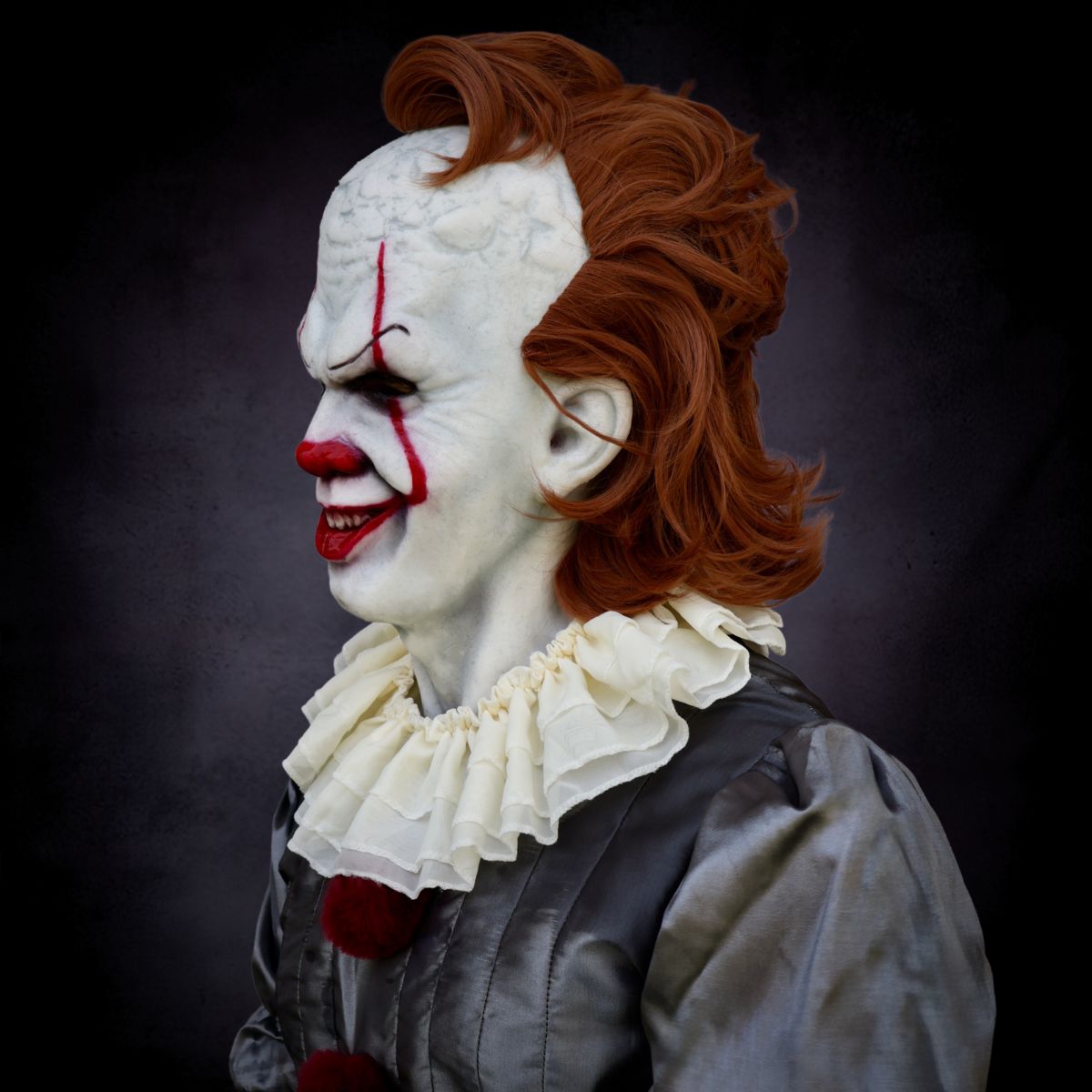 Pennywise Clown with white face, red hair, and ruffled collar full costume against a dark background