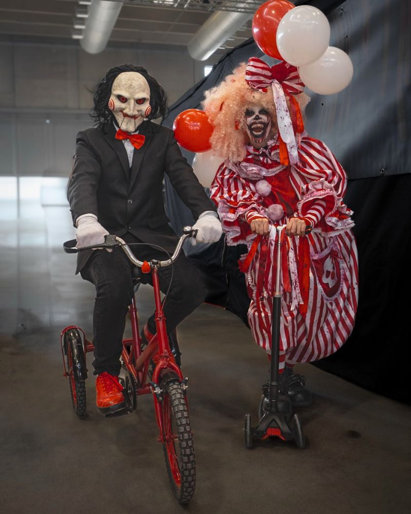 Customer wearing Billy the Puppet moving jaw mask riding a red tricycle next to a creepy clown in a red-and-white costume holding balloons — The Terror Lab.