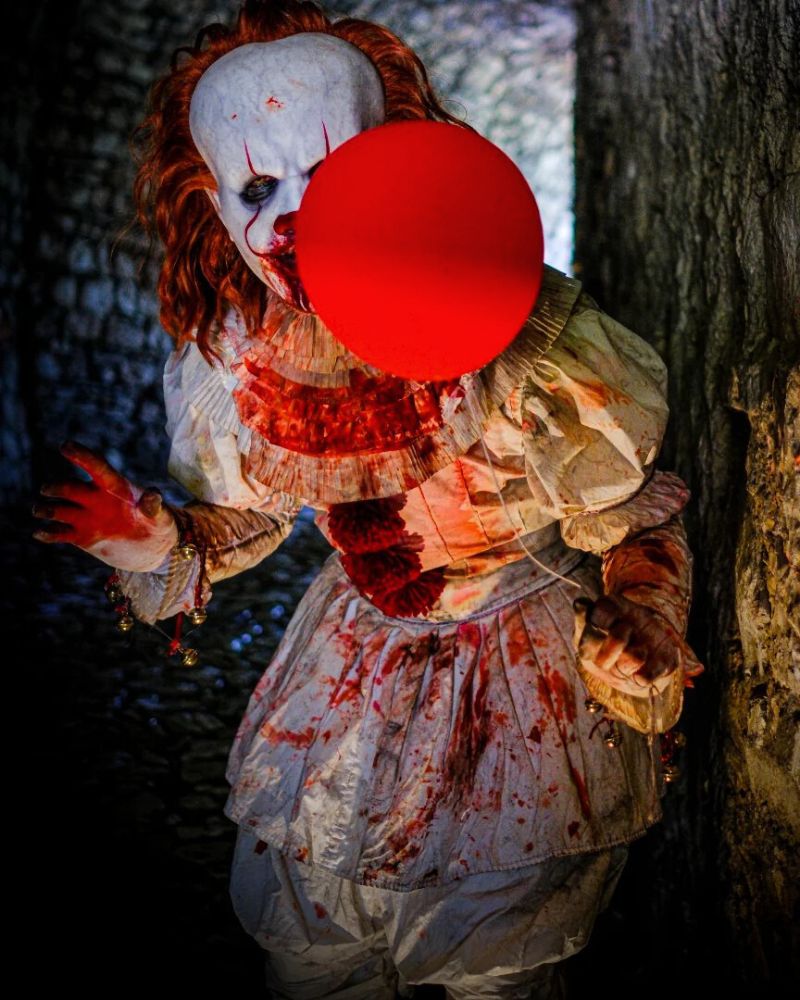 Pennywise clown full costume drenched in fake blood, lurking in a stone alley with red balloon obscuring part of the face — The Terror Lab
